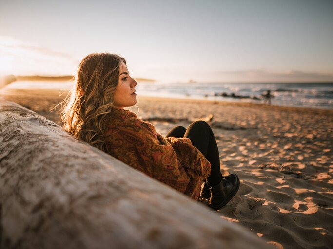 Frau sitzt nachdenklich am Strand bei Sonnenuntergang | © Getty Images/MarioGuti
