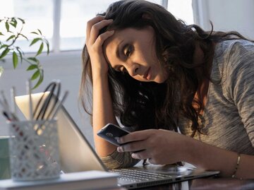 Junge Frau sitzt am Schreibtisch und schaut nachdenklich auf ihr Handy | © Getty Images/JGI/Tom Grill
