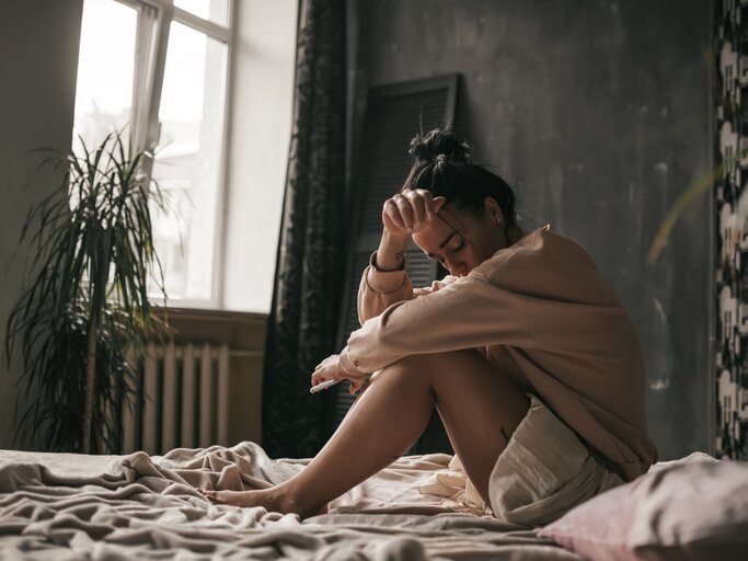 Junge Frau sitzt traurig und frustriert auf ihrem Bett | © Getty Images/Ol'ga Efimova/EyeEm