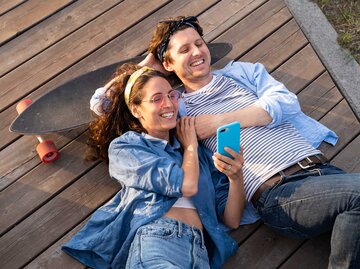 Ein junges befreundetes Paar liegt gemeinsam auf einem Holzsteg, lacht fröhlich und schaut ins Handy. | © Getty Images / Dmitry Marchenko / EyeEm