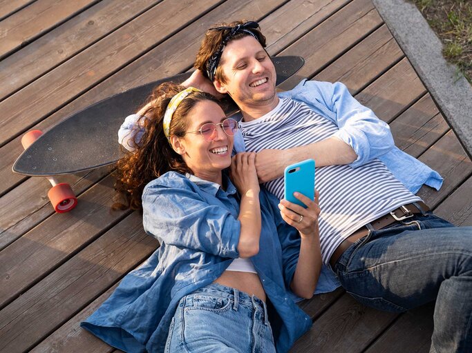 Ein junges befreundetes Paar liegt gemeinsam auf einem Holzsteg, lacht fröhlich und schaut ins Handy. | © Getty Images / Dmitry Marchenko / EyeEm