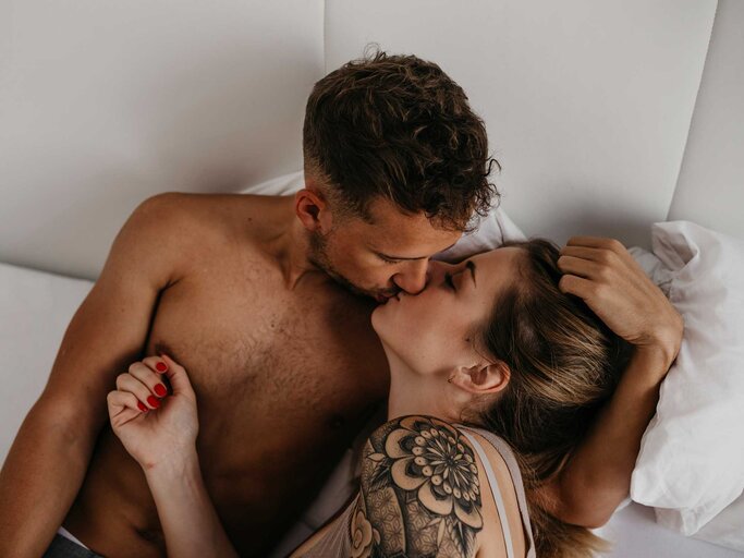 Frau liegt im Arm eines Mannes im Bett und küsst ihn.  | © Getty Images / Westend61