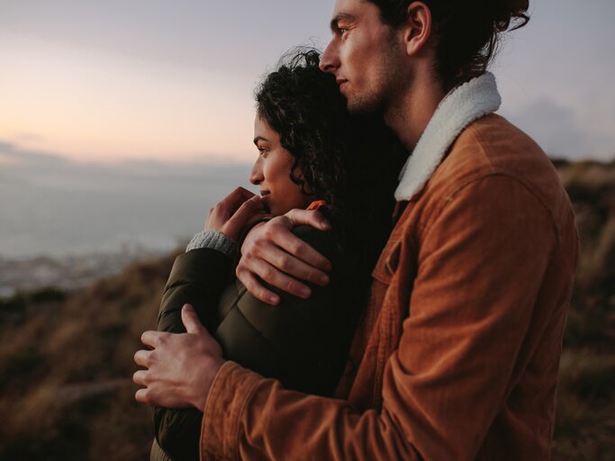 Mann und Frau stehen zusammen auf einem Berg und schauen in die Ferne während er sie von hinten umarmt | © Getty Images/jacoblund