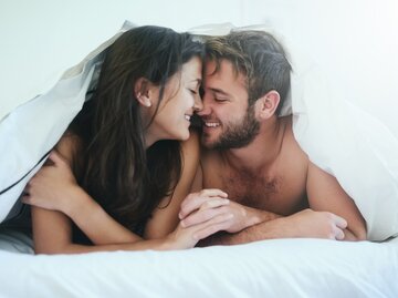 Junges Paar liegt zusammen im Bett unter der Bettdecke und lächelt sich an | © Getty Images/PeopleImages