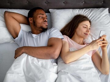 Frau liegt neben Mann im Bett und sieht auf ihr Handy | © Getty Images/Prostock-Studio