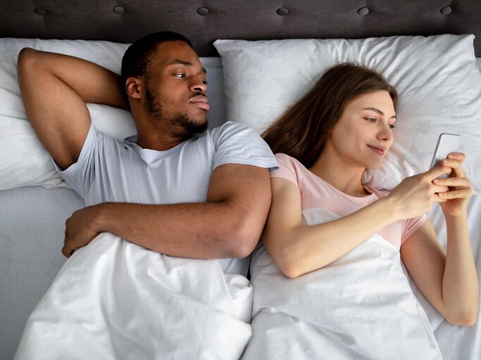 Frau liegt neben Mann im Bett und sieht auf ihr Handy | © Getty Images/Prostock-Studio