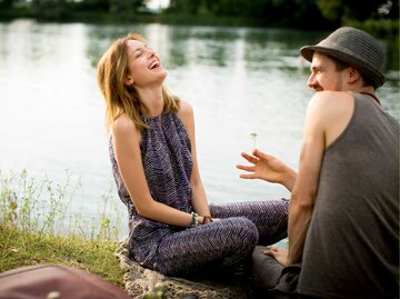Frau und Mann sitzen am See und lachen zusammen | © Getty Images/Oana Szekely