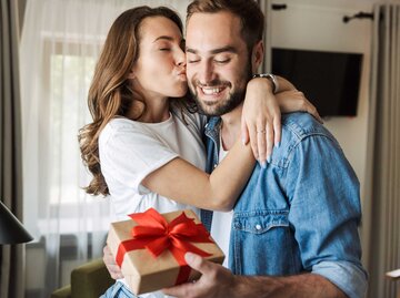 Frau küsst ihren Partner, der ein Valentinstagsgeschenk in der Hand hält | © Adobe Stock/Drobot Dean