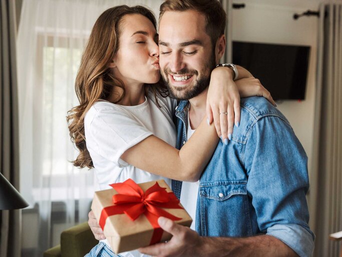Frau küsst ihren Partner, der ein Valentinstagsgeschenk in der Hand hält | © Adobe Stock/Drobot Dean