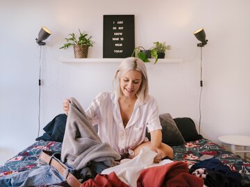 Eine junge Frau sitzt auf dem Bett und sortiert Kleidung aus | © GettyImages/beavera