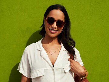 Frau mit Sonnenbrille steht vor einem grünen Hintergrund und lächelt | © Getty Images/PeopleImages