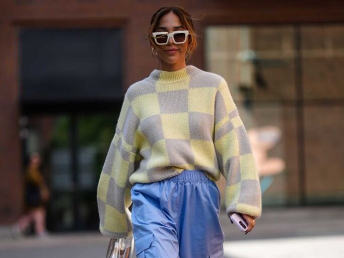 Frau trägt Sonnenbrille, blassgelben und blassgrauen Wollpullover mit Karomuster, eine blassblaue Cargohose mit weitem Bein aus Seidensatin | © Getty Images/Edward Berthelot