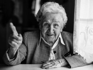 Alte Frau in kariertem Blazer sitzt mit erhobenem Zeigefinger am Tisch | © AdobeStock/De Visu