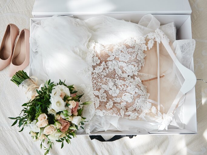 Hochzeitskleid ist in weißer Schachtel, beige Frauenschuhe und Brautstrauß liegen daneben. | © shutterstock/Mirage_studio
