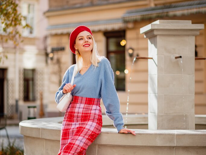 Modische glückliche, blonde Frau mit roter Mütze, blauem Kaschmirpullover und kariertem Rock schaut in die Ferne und lächelt. | © shutterstock/Victoria Chudinova