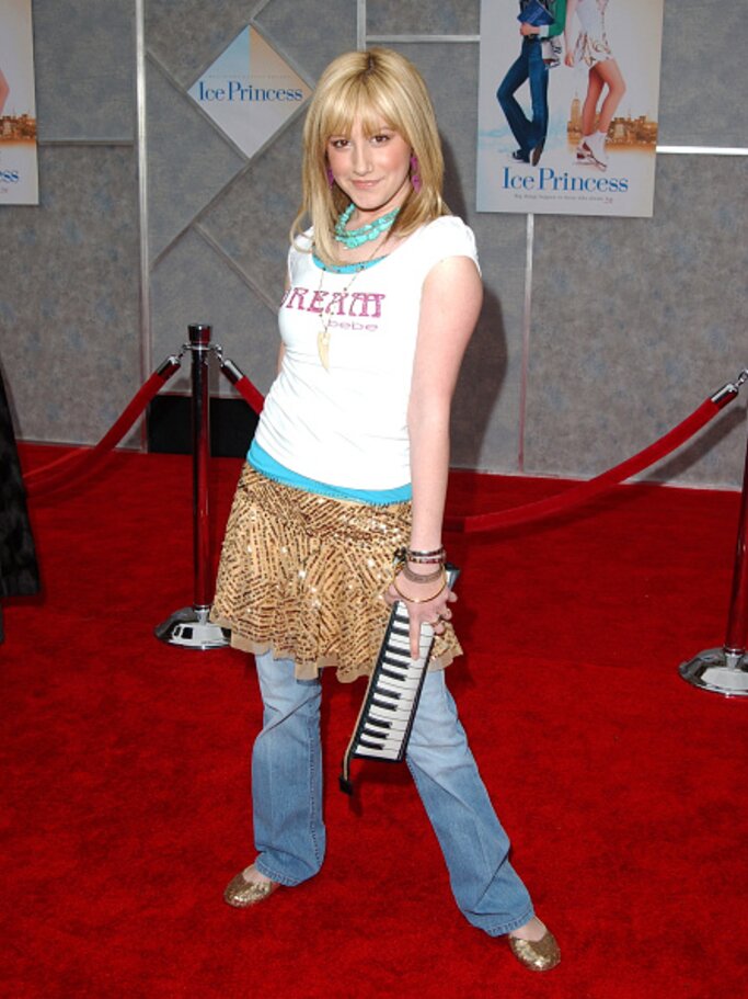 Ashley Tisdale trägt eine Jeans mit einem goldenen Rock darüber und ein weißes Shirt mit einer Aufschrift. Dazu trägt sie eine Tasche in Klaviertasten-Optik. | © Getty Images/ Jon Kopaloff 