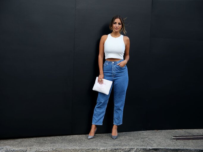 Junge Frau trägt weite Jeans und ein enges weißes Top ohne Ärmel. Dazu trägt sie eine weiße Clutch und graue Heels | © Getty Images/Don Arnold 