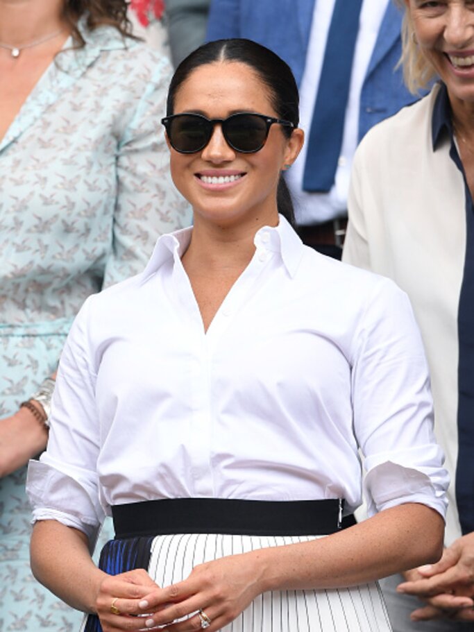 Herzogin Meghan trägt eine schwarze Sonnenbrille und eine weiße, hochgekrempelte Bluse mit einem blau-weißen Plisseerock. | © Getty Images/Karwai Tang 