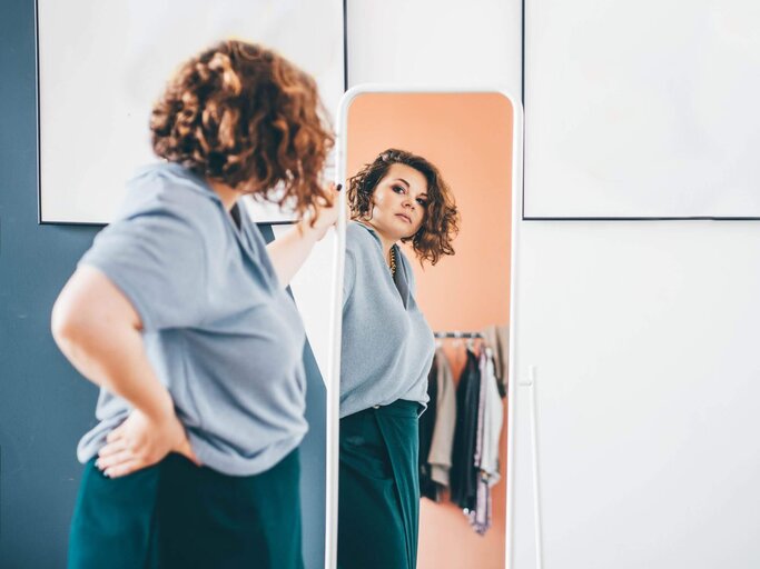 Frau schaut sich prüfend im Spiegel an | © Getty Images/Maria Korneeva