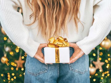 Frau mit langen Haaren und weißen Pullover steht vor einem Weihnachtsbaum und versteckt ein kleines Geschenk hinter ihrem Rücken. | © Getty Images / Yana Iskayeva