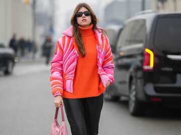 Frau trägt orangenen Pulli, pinke Daunenjacke, schwarze Hose, orangene Schuhe und pinke Handtasche | © Getty Images/Edward Berthelot 