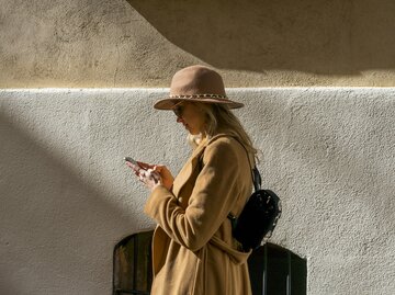Frau trägt beigen Mantel, beigen Hut, Sonnenbrille und schaut auf ihr Handy | © Getty Images/Westend61