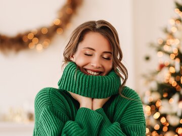 Frau kuschelt sich in ihren grünen Strickpullover ein und lächelt dabei | © Getty Images/Elizaveta Starkova