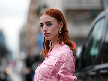 Eine rothaarige Frau trägt goldene Ohrringe von Designer Bottega Veneta und eine rosa Jeansjacke. | © Getty Images/Edward Berthelot 