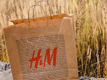 Eine Papiertüte von H&M mit dem roten H&M-Logo steht auf einem sommerlichen Feld. | © Adobe Stock/Татьяна