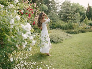 Junge Frau in langem, weißen Kleid vor blühenden Rosen im Garten | © AdobeStock/polinaloves
