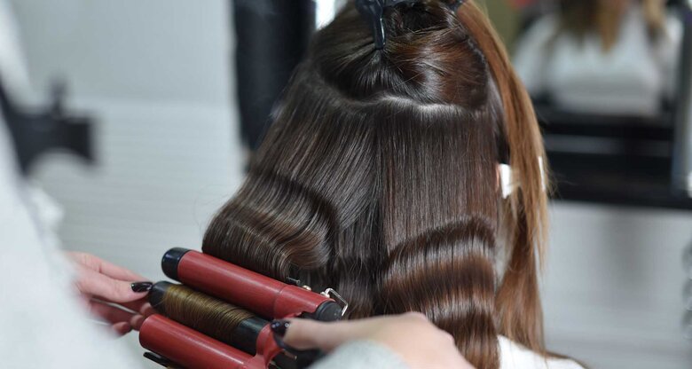 Frau sitzt beim Friseur und bekommt mit einem Welleneisen locken in die Haare gestylt. | © Getty Images / Predrag Popovski