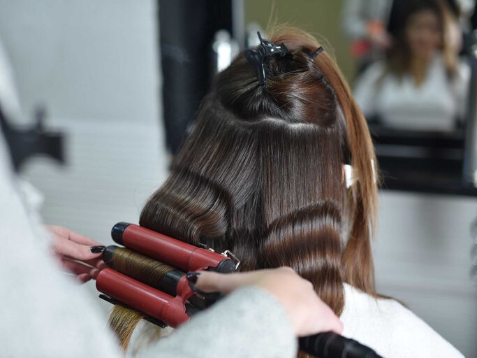 Frau sitzt beim Friseur und bekommt mit einem Welleneisen locken in die Haare gestylt. | © Getty Images / Predrag Popovski