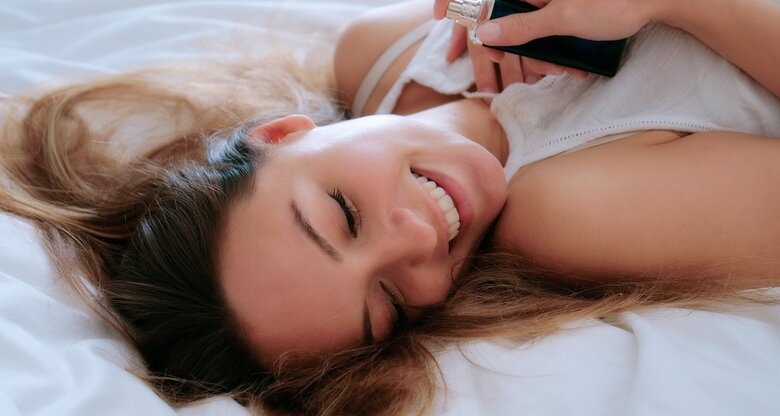 Eine junge Frau liegt lächelnd auf dem Bett und hält ein Pheromone Parfum an ihren Hals | © GettyImages/John Slater