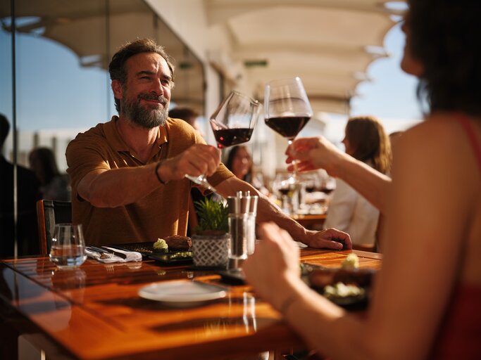 Reifer Mann in braunem Poloshirt beim fröhlichen Anstoßen mit einer Frau auf sonniger Restaurantterrasse mit elegantem Flair. | © AIDA