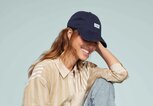 Junge Frau mit welligem braunem Haar lacht entspannt, trägt eine beige gestreifte Bluse, Jeans, weiße Sneaker und eine dunkle Baseballkappe. | © TOMS