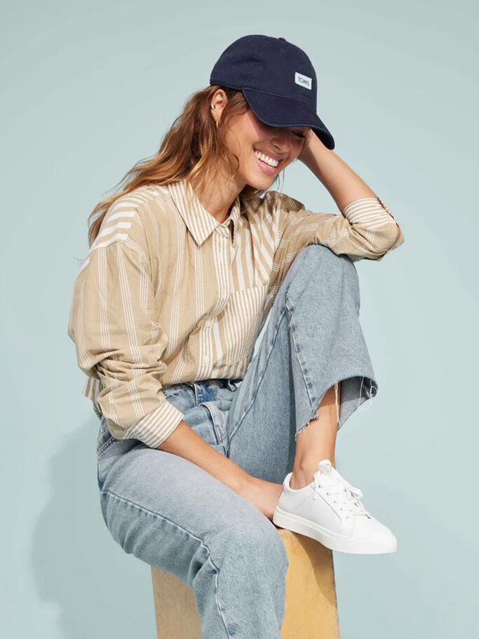 Junge Frau mit welligem braunem Haar lacht entspannt, trägt eine beige gestreifte Bluse, Jeans, weiße Sneaker und eine dunkle Baseballkappe. | © TOMS