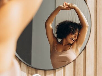 Zufriedene junge Frau mit Afro hebt lächelnd die Arme vor einem runden Spiegel an, Sonnenlicht fällt auf helle Holzvertäfelung. | © stock.adobe.com/Maria Vitkovska