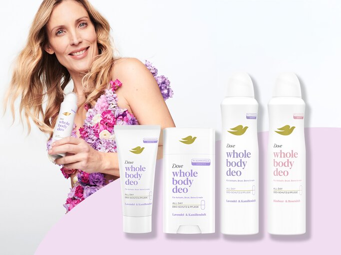 Frau mit welligem, blondem Haar trägt Kleid aus rosa und lila Blüten und präsentiert Dove Whole Body Deo mit sanftem Lächeln. | © Dove / myself.de