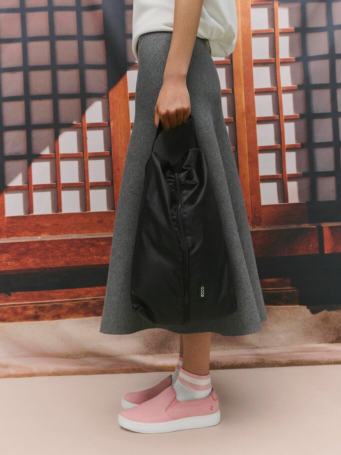 Eine Frau in grauem Midirock und weißem Oberteil hält eine schwarze Tasche, dazu trägt sie rosa Sneaker und gestreifte Socken. | © ECCO