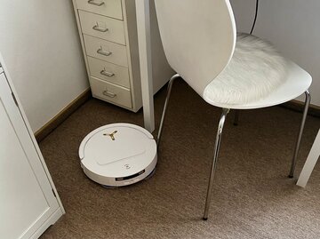 Ein weißer Roboterstaubsauger fährt auf einem hellbraunen Teppich unter einem modernen Stuhl mit weißem Fellkissen. | © myself