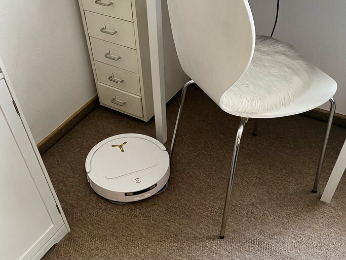 Ein weißer Roboterstaubsauger fährt auf einem hellbraunen Teppich unter einem modernen Stuhl mit weißem Fellkissen. | © myself