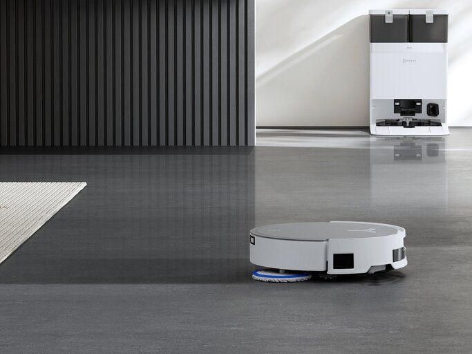 Ein moderner Saug- und Wischroboter reinigt einen glänzenden Betonboden, im Hintergrund steht eine weiße Ladestation an der Wand. | © ECOVACS