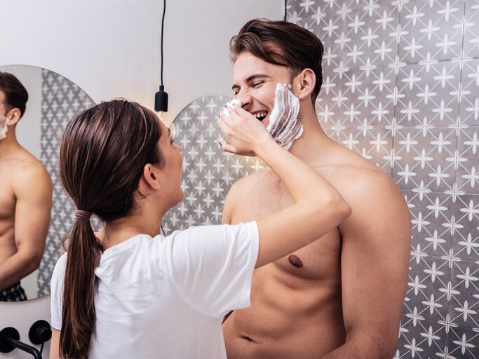 Junge Frau mit braunem Pferdeschwanz trägt lachend Rasierschaum auf das Gesicht ihres freundlichen, muskulösen Partners im Bad auf. | © stock.adobe.com/Viacheslav Yakobchuk
