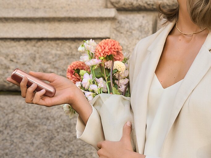 Junge Frau in weißem Blazer und goldener Halskette hält einen Blumenstrauß und ein roségoldenes Accessoire vor Steinwand. | © Ploom