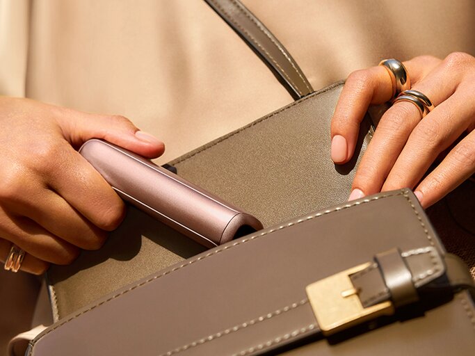 Nahaufnahme von zwei Händen mit gepflegten Nägeln und Schmuck, die ein metallisch glänzendes Accessoire aus einer taupefarbenen Tasche holen. | © Ploom