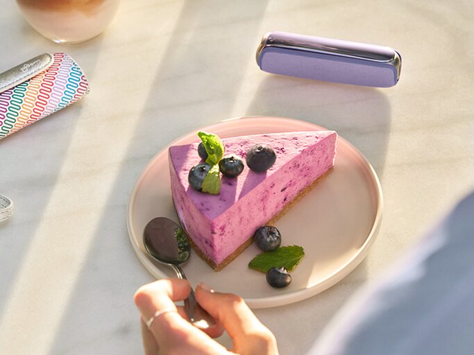 Hand mit silbernem Ring greift zu einem Löffel neben Blaubeerkuchen, bunte Serviette und violett-glänzendes Accessoire im Hintergrund. | © Ploom