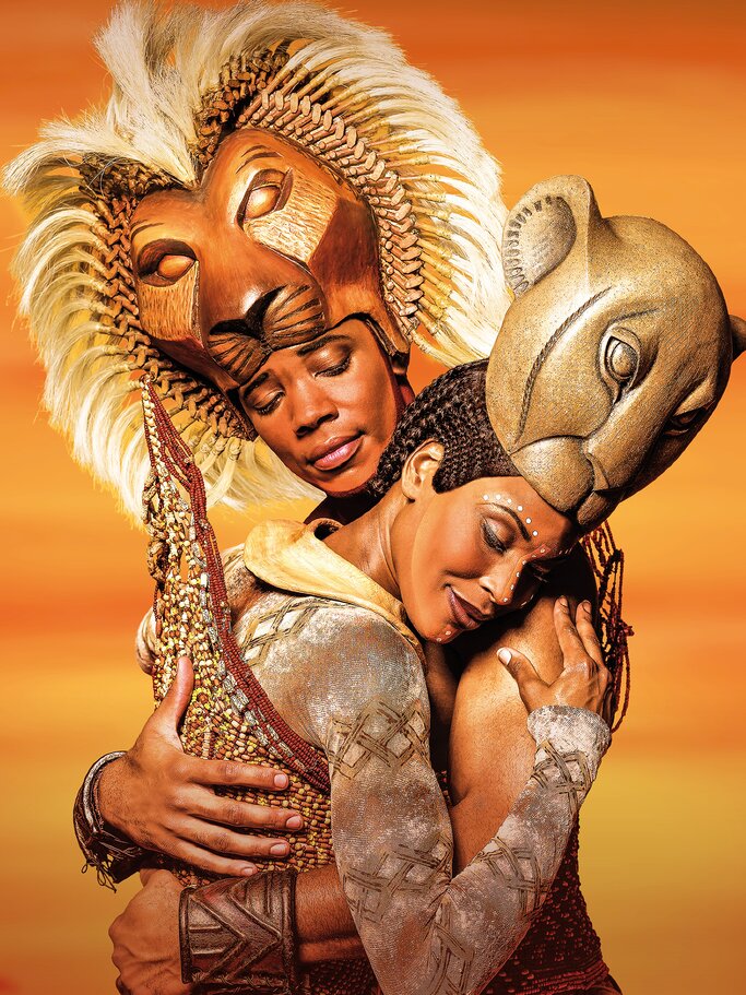Zwei Darsteller in kunstvollen Löwenmasken und afrikanisch inspirierten Kostümen umarmen sich innig vor warmem, orangefarbenem Hintergrund. | © Lois Greenfield
