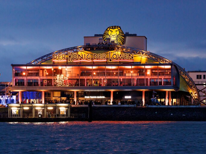 Das hell erleuchtete Stage Theater im Hafen Hamburgs mit dem leuchtenden Schriftzug Der König der Löwen vor dunkelblauem Abendhimmel. | © Morris Mac Matzen/mmacm.com