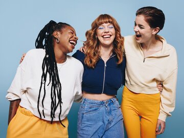 Drei junge Frauen in bunten Outfits lachen herzlich und umarmen sich vor einem hellblauen Hintergrund. | © Jacob Lund/stock.adobe.com