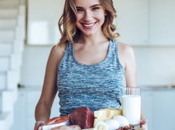 Frau in sportlichem Outfit hält ein Schneidebrett mit eiweißreichen Nahrungsmitteln wie gekochtem Ei, Kuhmilch, Käse, ein Lachssteak und rohes Fleisch | © iStock / Vasyl Dolmatov